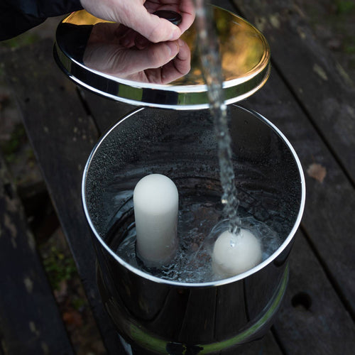 Hand lifting the polished stainless steel lid of a mid-size gravity water filter as fresh water pours inside toward two ceramic filter elements, suitable for farm, garden, and camping use.