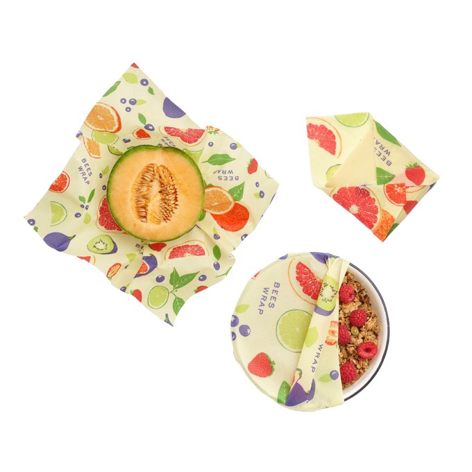 Three-piece set of handcrafted beeswax wraps with colorful fruit and leaf patterns, used for covering melon half, wrapped snack, and bowl of granola and berries, USA Made, Kitchen & Food and Storage and Serving collections.