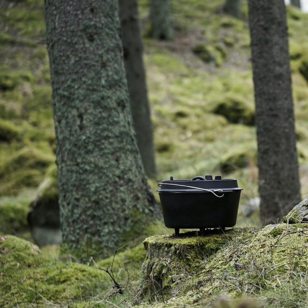 Cast iron Dutch oven with legs resting on a moss-covered tree stump in a forest setting, highlighting durability and outdoor cooking versatility for Kitchen & Food, Cookware, Cast Iron, Best Sellers, Promo Eligible collections.