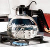 Clear borosilicate glass teakettle with black handle and lid boiling on a gas stove, designed for Kitchen & Food and Brewing Coffee and Tea collections.
