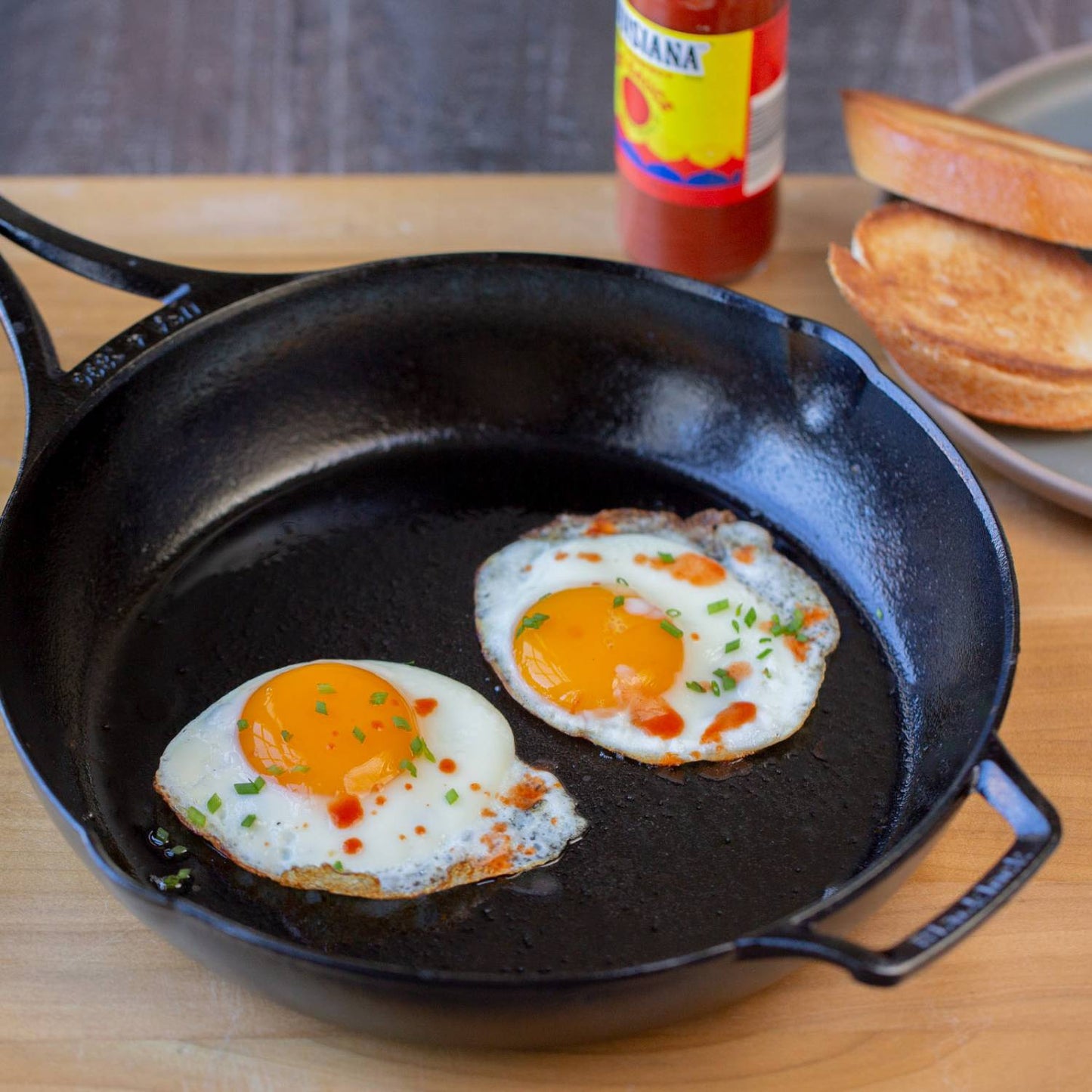 Handcrafted lightweight cast iron skillet made in USA with two sunny-side-up eggs garnished with chives and hot sauce, shown alongside toasted bread for Kitchen & Food and Cookware collections.