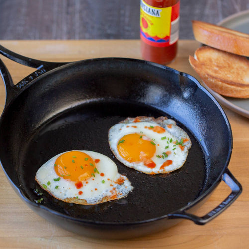 Handcrafted lightweight cast iron skillet made in USA with two sunny-side-up eggs garnished with chives and hot sauce, shown alongside toasted bread for Kitchen & Food and Cookware collections.