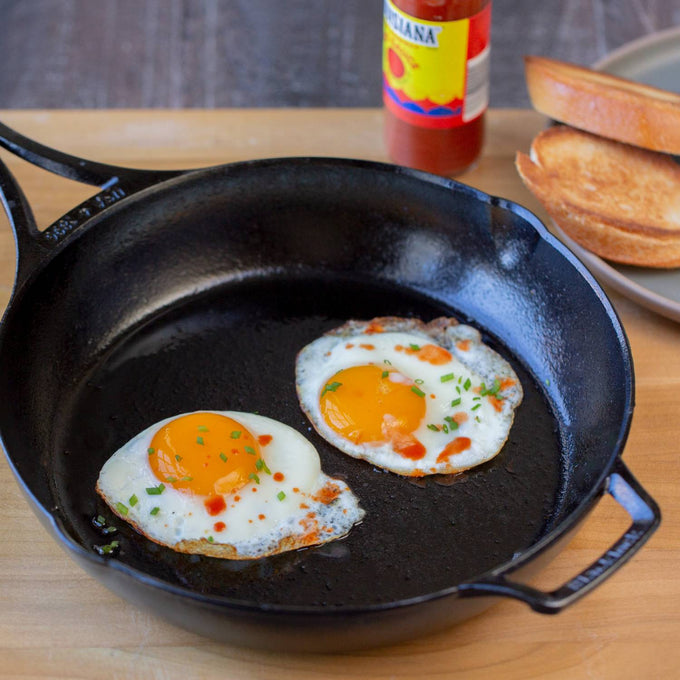 Handcrafted lightweight cast iron skillet made in USA with two sunny-side-up eggs garnished with chives and hot sauce, shown alongside toasted bread for Kitchen & Food and Cookware collections.