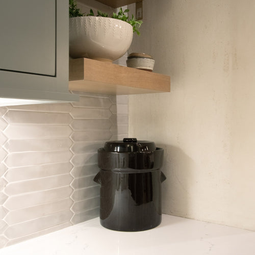 Handcrafted black stoneware fermenting crock with lid, placed on a white kitchen countertop under a wooden shelf, ideal for Kitchen & Food, Food Preservation, Fermenting and Pickling, Best Sellers, and Promo Eligible collections.
