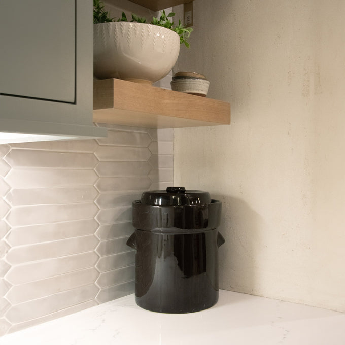 Handcrafted black stoneware fermenting crock with lid, placed on a white kitchen countertop under a wooden shelf, ideal for Kitchen & Food, Food Preservation, Fermenting and Pickling, Best Sellers, and Promo Eligible collections.