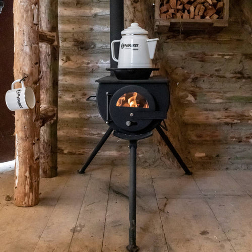 Compact black wood-burning stove with visible flames inside, supporting a white enamel kettle on top, set in a rustic cabin with hanging mug and stacked firewood, ideal for camping and outdoor use.