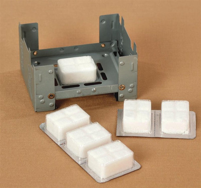 Compact fold-out metal camping stove with white solid fuel cubes arranged on and beside it, designed for outdoor use in camping, emergency supplies, and garden tool collections