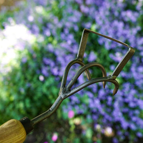 Close-up of a handcrafted cultivator and weeder garden tool featuring a durable steel head and smooth ash wood handle, set against a backdrop of purple flowers in a garden setting.