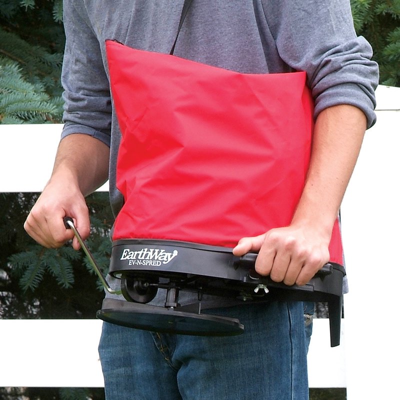 Person holding a red nylon shoulder spreader with a black base and crank handle, designed for evenly dispersing seeds or granular material, part of Farm, Garden & Tool, Seeds and Planting, USA Made, Best Sellers, and Promo Eligible collections.