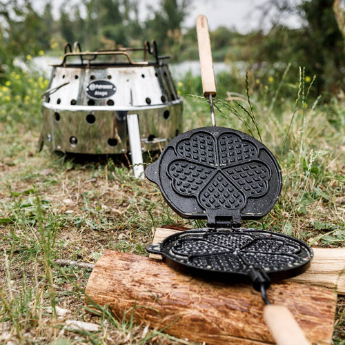 Handcrafted cast iron rotating waffle iron with wooden handles and heart-shaped molds, set on a log outdoors near a portable camping stove, ideal for Farm, Garden & Tool, Camping, and Best Sellers collections.