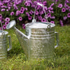 Large galvanized steel watering can with a seamless rust-resistant finish and sturdy handle, placed on green grass with purple flowers in the background, ideal for farm and garden use.