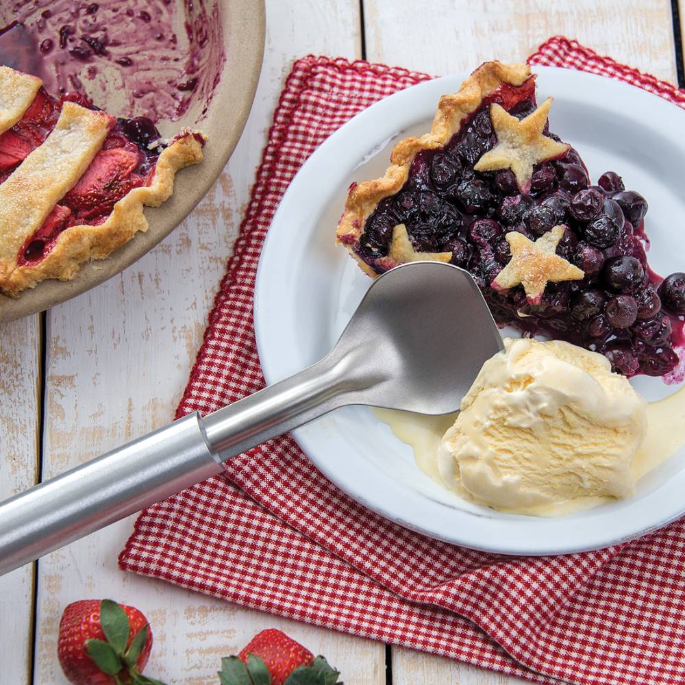 Handcrafted stainless steel ice cream scoop with a stub-nose end resting beside a slice of star-topped berry pie and melting vanilla ice cream on a white plate, featured in Kitchen & Food, Specialty Equipment, Making Ice Cream, Storage and Serving, USA Made, and Best Sellers collections.