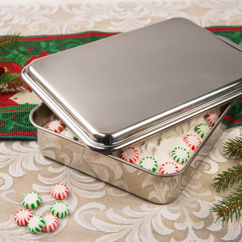 Rectangular stainless steel cake pan with a raised lid slightly ajar, revealing red and green peppermint candies inside, set on a festive holiday tablecloth.