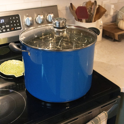 Glossy blue 21.5-quart water bath canner with a clear glass lid and black handles, sitting on a stovetop in a kitchen setting with utensils and countertop accessories in the background.