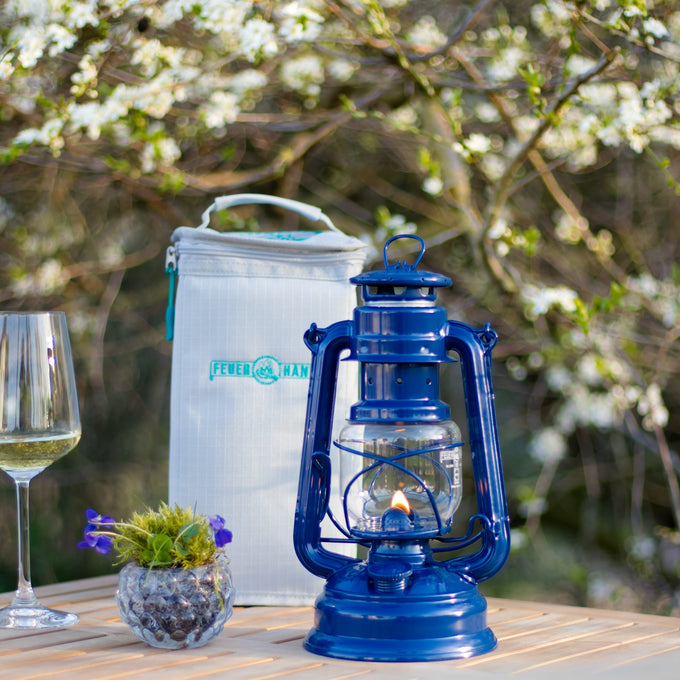 Handcrafted deep blue galvanized oil lantern with thermal heat-resistant SUPRAX globe, glowing warmly on a wooden table alongside a wine glass and cooler bag, perfect for camping, emergency supplies, and outdoor lighting from best sellers and promo collections.