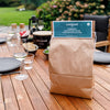 Brown paper bag of charcoal placed on a wooden outdoor table set for a casual barbecue, surrounded by plates, glassware, and seating in a garden setting, ideal for grilling enthusiasts.