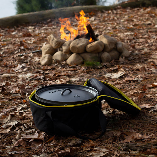 Black weather-resistant tote with bright yellow piping and thick padding, open to reveal cast iron cookware inside, set on forest floor near a stone-lined campfire, ideal for camping and outdoor cookware storage in Farm, Garden & Tool and Kitchen & Food collections.