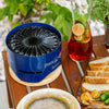 Compact blue tabletop charcoal grill with unique spiral top design resting on a wooden trivet, set on an outdoor wooden table with sliced bread and a glass pitcher of fruit-infused iced tea, featured in Farm, Garden & Tool and Grilling collections.