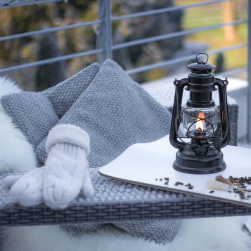 Handcrafted black galvanized oil lantern with thermal heat-resistant SUPRAX globe glowing warmly on an outdoor wicker bench with cozy knit pillows and white mittens, ideal for Lamps & Lighting, Camping, and Emergency Supplies.