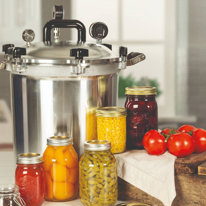 Stainless steel pressure canner next to an assortment of sealed jars filled with preserved fruits and vegetables, alongside fresh tomatoes on a kitchen counter, highlighting food preservation essentials.
