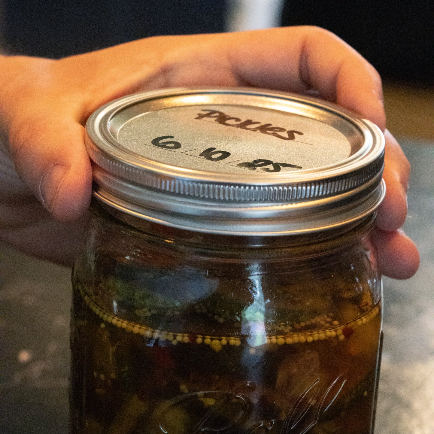 Hand holding a wide-mouth glass jar filled with pickles sealed with metal canning lid and band featuring BPA-free sealing compound, ideal for Kitchen & Food and Food Preservation collections.