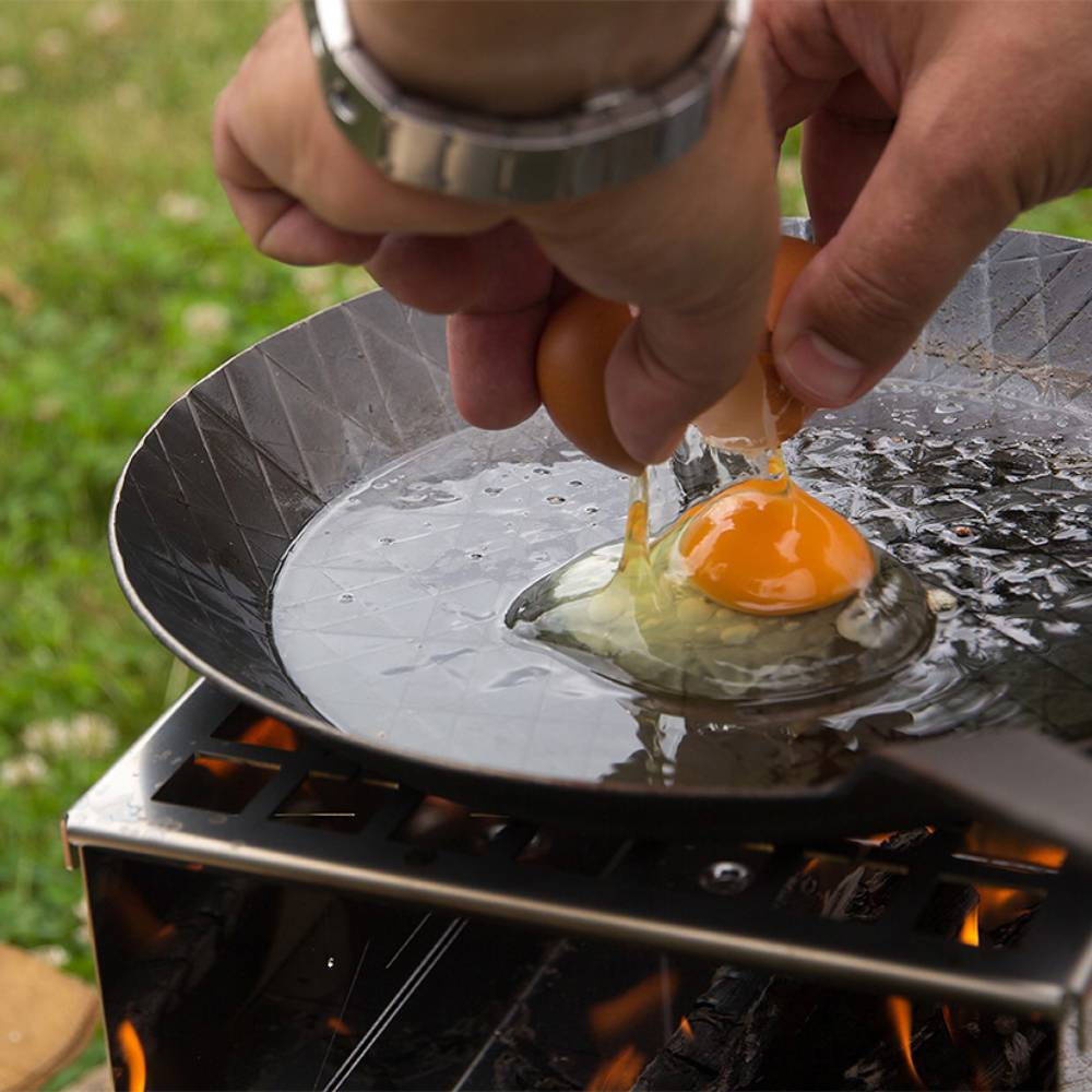 Handcrafted wrought iron pan heating over a portable outdoor stove with a fresh egg being cracked for cooking, ideal for Farm, Garden & Tool, Camping, Kitchen & Food, Cookware, Pots and Pans, Best Sellers, Promo Eligible collections.