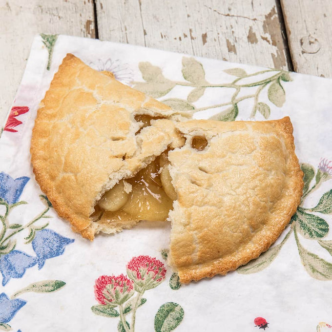 Golden-brown handmade fruit turnover with a flaky crust revealing a sweet apple filling, presented on a floral cloth, highlighting a handcrafted treat from Kitchen & Food and Snacks and Sweets collections.