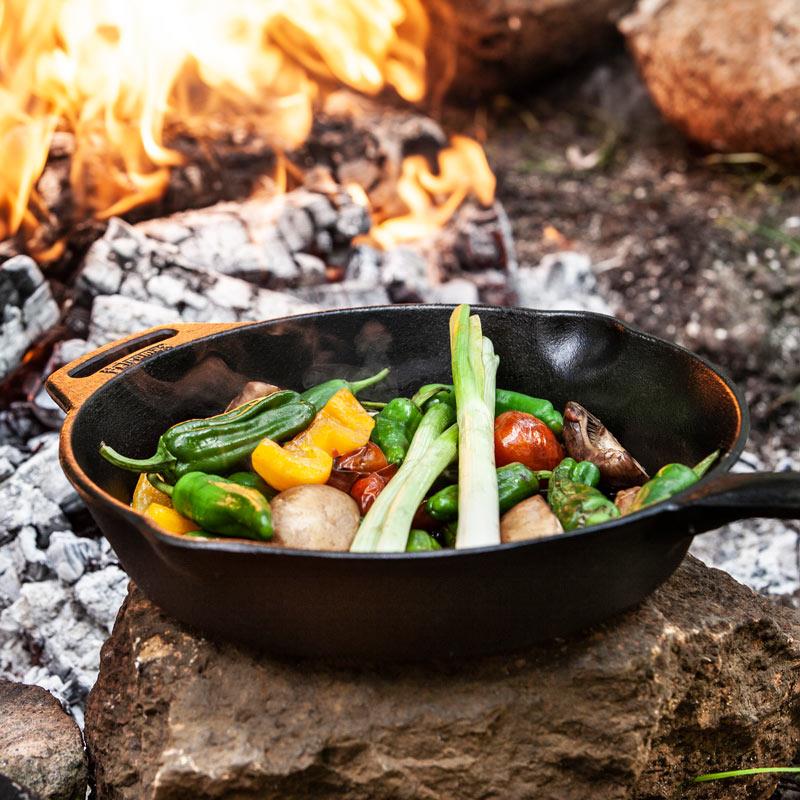 Cast iron skillet with long handle cooking a colorful mix of fresh vegetables beside an open campfire on a rock surface, ideal for Farm, Garden & Tool, Kitchen & Food, Camping, Cookware, Cast Iron, Best Sellers, and Promo Eligible collections.