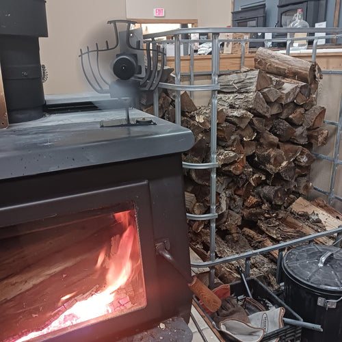 Rotating heat-powered stove fan on a wood-burning stove with visible flames, beside a metal rack stacked with firewood, ideal for stoves and hearth accessories collections.