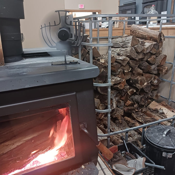Rotating heat-powered stove fan on a wood-burning stove with visible flames, beside a metal rack stacked with firewood, ideal for stoves and hearth accessories collections.