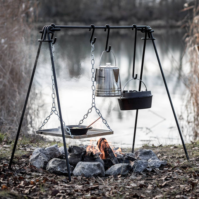Black-coated steel fire bridge with adjustable hooks and chain-suspended kettle, Dutch oven, and grill grate over a stone-lined campfire, perfect for camping, grilling, and Farm, Garden & Tool collections.