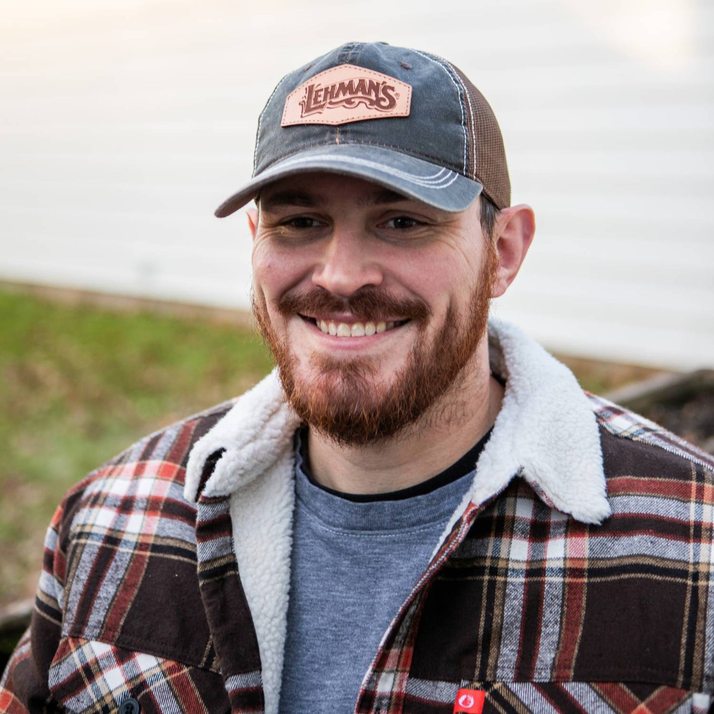 Smiling man wearing an adjustable Lehman's leather patch hat with weathered cotton front and breathable mesh back, styled with a plaid jacket in Health & Wellness and Clothing collections.