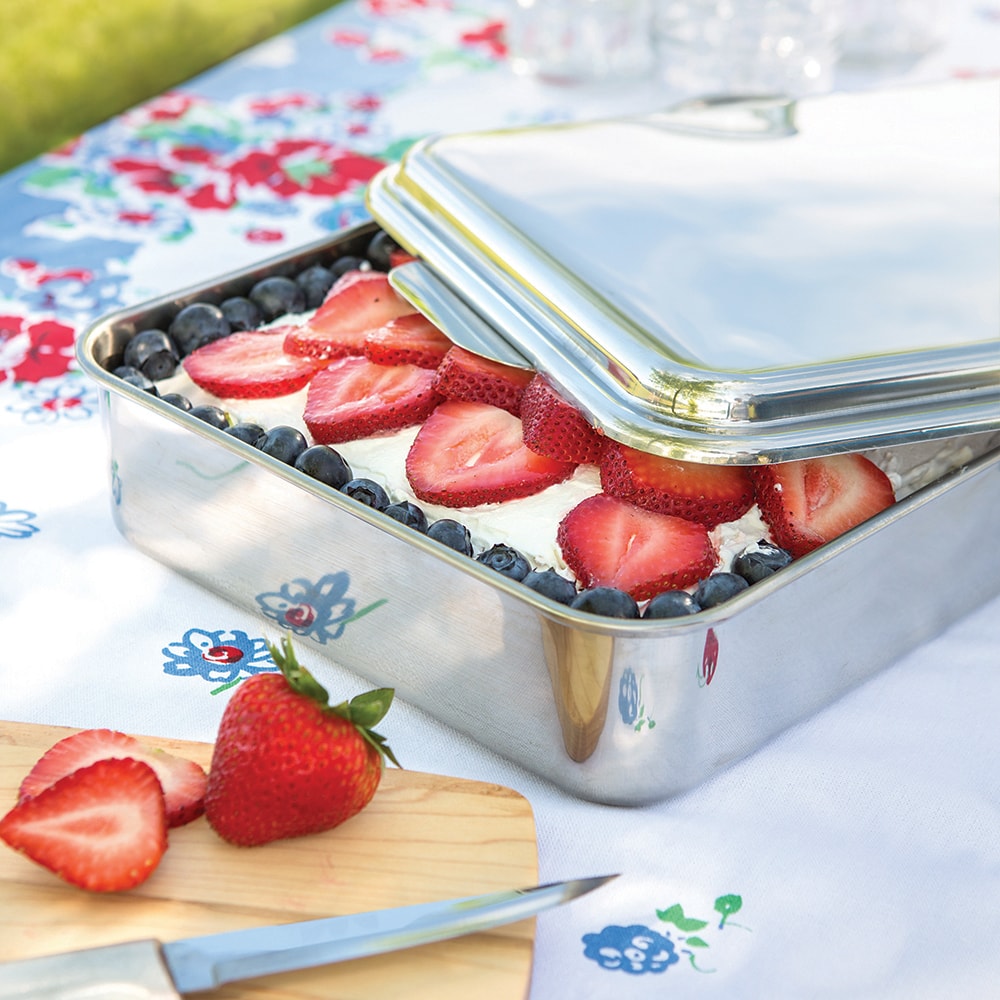 Rectangular stainless steel cake pan with lid partially open, showing a dessert topped with fresh sliced strawberries and blueberries, set on a floral tablecloth in a bright kitchen setting.