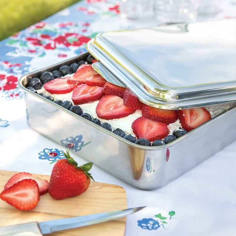 Rectangular stainless steel cake pan with lid partially open, showing a dessert topped with fresh sliced strawberries and blueberries, set on a floral tablecloth in a bright kitchen setting.