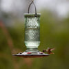 Clear vintage-inspired glass hummingbird feeder with etched decorative patterns, five feeding ports, and burnt penny plastic base, hanging outdoors in a garden setting.