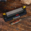 Angle-guided benchtop knife sharpener with coarse and fine abrasive surfaces in black base with yellow adjustment knobs, shown with a pocket knife on a rustic wooden workbench, suitable for Farm, Garden & Tool and Kitchen & Food collections.