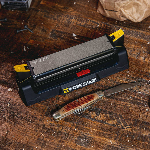 Angle-guided benchtop knife sharpener with coarse and fine abrasive surfaces in black base with yellow adjustment knobs, shown with a pocket knife on a rustic wooden workbench, suitable for Farm, Garden & Tool and Kitchen & Food collections.