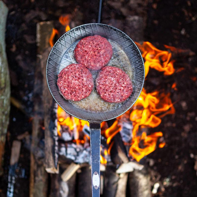 Petromax wrought iron pan with a long handle cooking three raw burger patties over an open wood fire in an outdoor campsite, perfect for Farm, Garden & Tool, Kitchen & Food, Camping, Cookware, Pots and Pans, Best Sellers, and Promo Eligible collections.