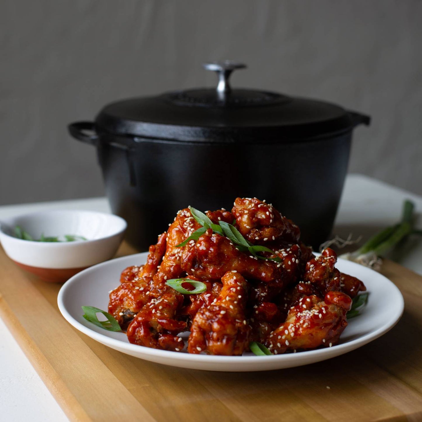 Lightweight black cast iron Dutch oven with lid behind a white plate of glazed chicken wings garnished with sesame seeds and sliced green onions, ideal for Kitchen & Food, Cookware, Cast Iron, USA Made, Best Sellers, Promo Eligible collections.