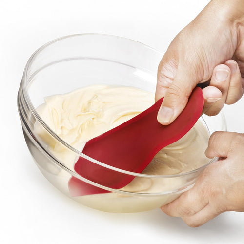 Red flexible silicone scraper with a thin metal core scraping batter from a clear glass bowl, designed for easy cleanup and smooth mixing in Kitchen & Food and Baking Supplies collections.