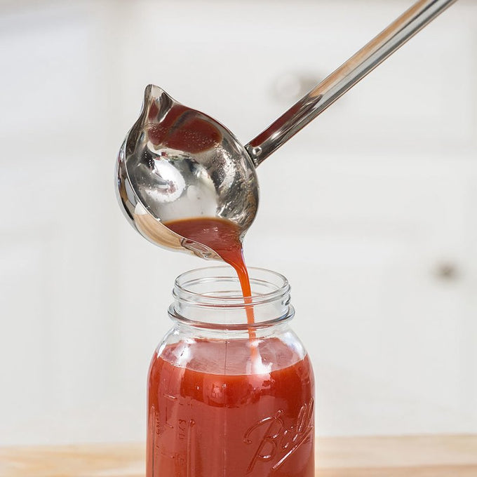 Shiny stainless steel double-spout ladle pouring red liquid into a clear glass jar, ideal for Kitchen & Food, Farm, Garden & Tool, Storage and Serving, Best Sellers, and Homesteading collections.