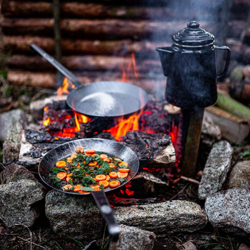 Handcrafted wrought iron pan with long handle cooking sliced carrots and greens over an open wood fire, accompanied by a black metal kettle, perfect for Farm, Garden & Tool, Kitchen & Food, Camping, Cookware, Pots and Pans, Best Sellers, and Promo Eligible collections.
