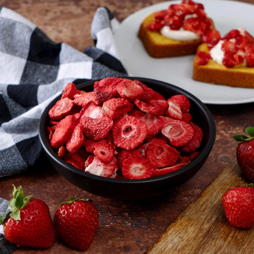 Black bowl filled with vibrant red freeze-dried strawberry slices, surrounded by fresh strawberries, a black and white checkered cloth, and two dessert slices topped with strawberries and cream, featured for kitchen and emergency supplies collections.