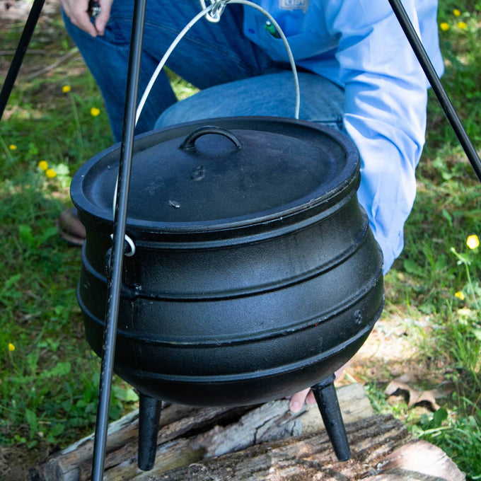 Handcrafted cast iron campfire kettle with a ribbed rounded body, three sturdy legs, and wire bail handle, set outdoors on wood with person adjusting it, ideal for Farm, Garden & Tool, Camping, Best Sellers, and Promo Eligible collections.
