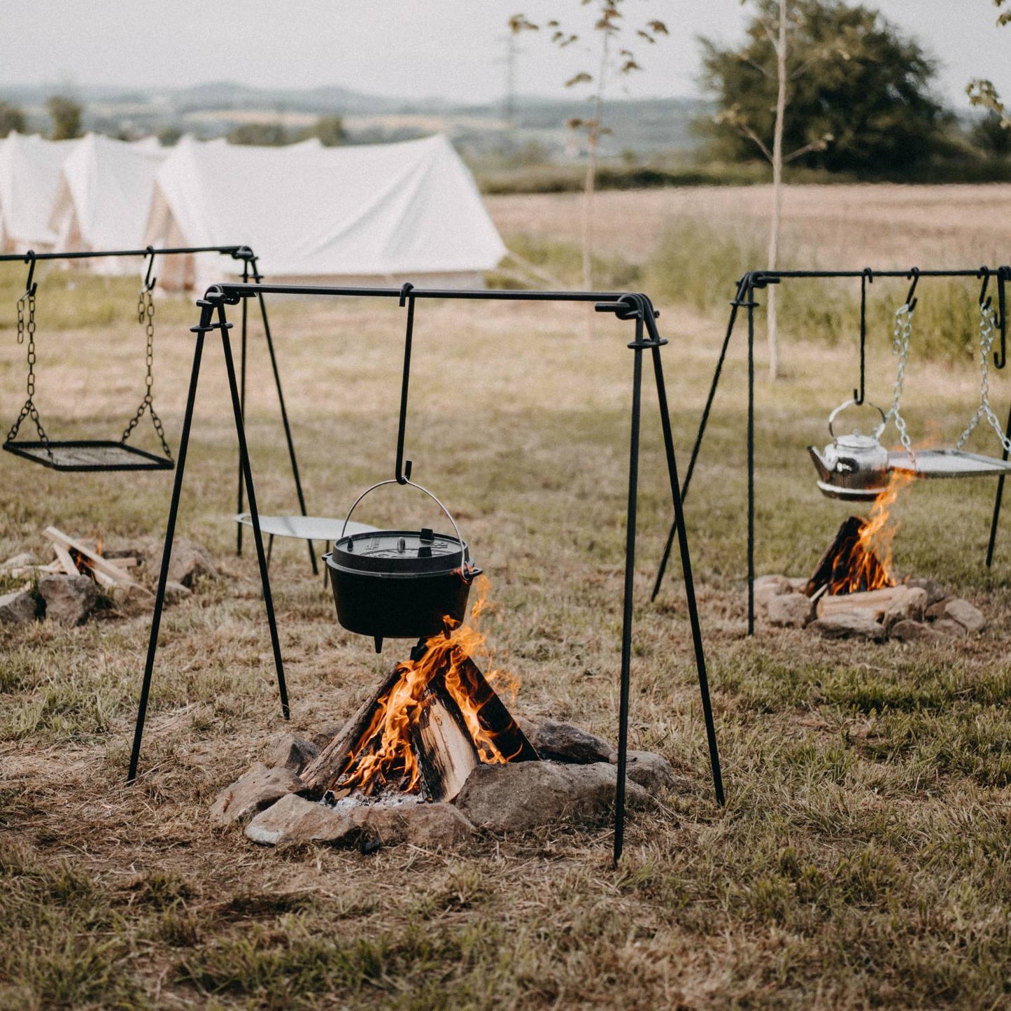 Outdoor cooking setup featuring sturdy black-coated metal fire bridges with four-leg stability and chain-hung hooks, holding pots and grills over campfire pits in a grassy field, perfect for camping and grilling in Farm, Garden & Tool and Stoves collections.