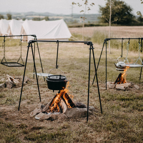 Outdoor cooking setup featuring sturdy black-coated metal fire bridges with four-leg stability and chain-hung hooks, holding pots and grills over campfire pits in a grassy field, perfect for camping and grilling in Farm, Garden & Tool and Stoves collections.