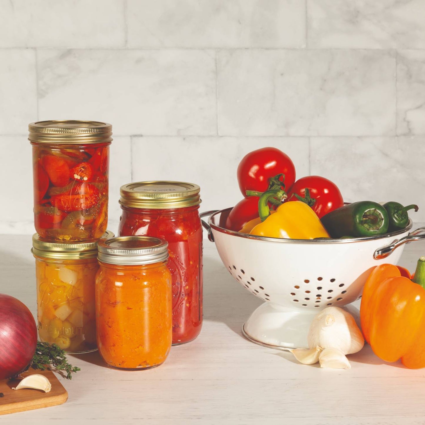 Four glass jars filled with preserved vegetables and sauces sealed with wide mouth metal lids, next to fresh bell peppers, tomatoes, garlic, and an onion in a white colander, highlighting Kitchen & Food and Food Preservation collections.