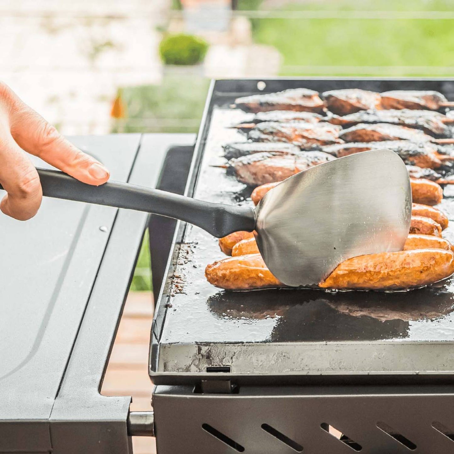 Flexible stainless steel spatula with a large flat blade and black handle lifting grilled sausages on an outdoor flat-top grill, ideal for Kitchen & Food and Grilling collections.