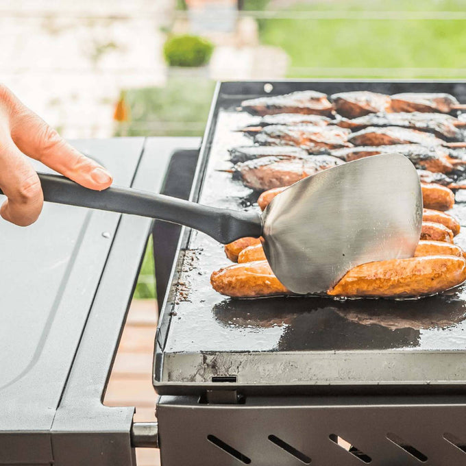Flexible stainless steel spatula with a large flat blade and black handle lifting grilled sausages on an outdoor flat-top grill, ideal for Kitchen & Food and Grilling collections.