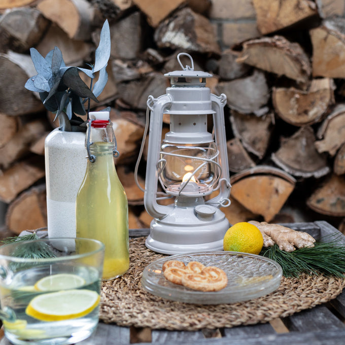 Handcrafted white galvanized oil lantern with thermal heat-resistant SUPRAX globe softly glowing on a rustic woven mat surrounded by citrus, ginger, and natural decor, perfect for Camping, Lamps & Lighting, and Emergency Supplies collections.